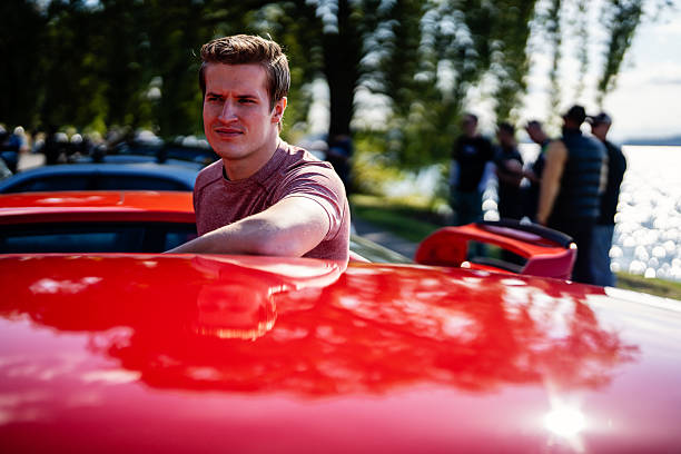A man stands by his red muscle car at a meetup at a park along the lake.  There are other cars in the image.  This was taken at a Sunday meetup along Lake Washington in Seattle Washington State.
