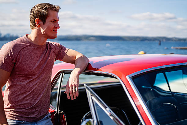 A man stands by his red muscle car at a meetup at a park along the lake.  This was taken at a Sunday meetup along Lake Washington in Seattle Washington State.