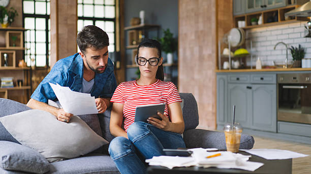 istock Couple calculating bills at home using tablet and calculator. Young couple working on computer while calculating finances sitting on couch. Young man with wife at home analyzing their finance with documents. 2240546590