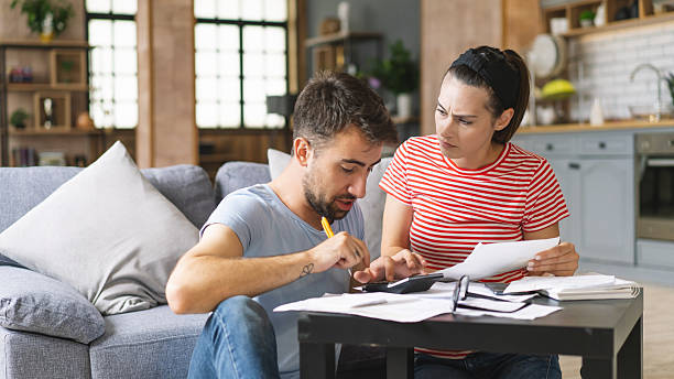 istock Couple calculating bills at home using calculator. Couple checking bills and documents while calculating finances sitting in the living room. Man and woman at home analyzing their finance with documents. 2240554656