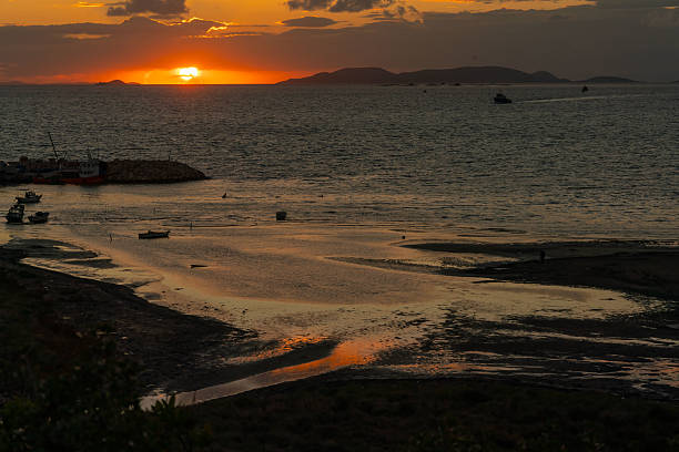 At sunset in Didyma (Didim), small boats rest on calm shallow waters reflecting the orange and pink tones of the sky. A distant ship appears on the horizon, while soft light glows across the tranquil seascape.