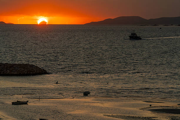 At sunset in Didyma (Didim), small boats rest on calm shallow waters reflecting the orange and pink tones of the sky. A distant ship appears on the horizon, while soft light glows across the tranquil seascape.