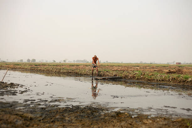 A solitary man stands in a shallow, muddy ditch within a vast rural field, his reflection mirrored in the water. The scene conveys hard work, resilience, and a connection to the land in a quiet countryside.