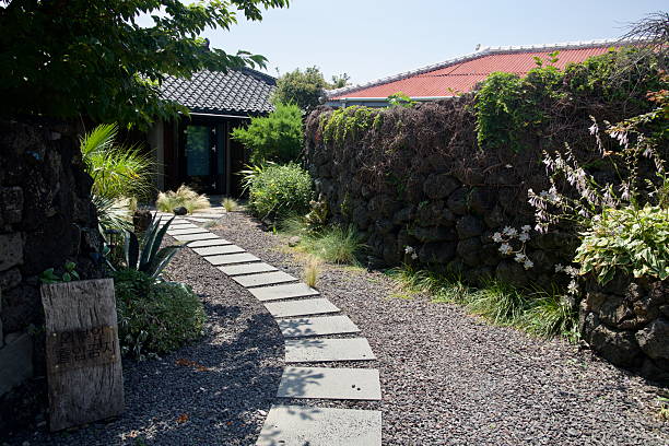 In Gimnyeong's Metal Craft Mural Village, a steppingstone path curves through a planted courtyard beside lavastone walls toward a renovated house entrance, with a small wooden notice at the edge of the walk.