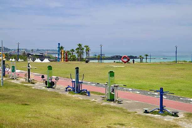 Outdoor fitness machines line a coastal park above Hamdeok Seoubong Beach, with public art, tents and the blue bay beyond the basalt breakwaters on Jeju's northeast coast.