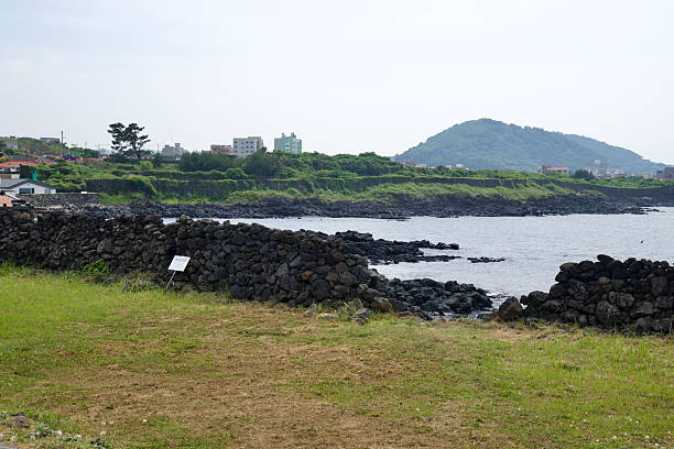 Basalt field walls and black rock ledges edge the Samyang coast with Sarabong rising beyond a green seawall, seen across calm inshore waters.