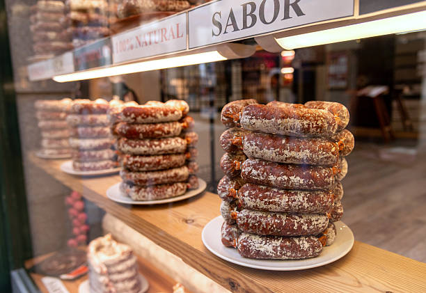 Stacks of Spanish cured sausage, dusted with white mold, are neatly displayed on a wooden shelf. The food is labeled \
