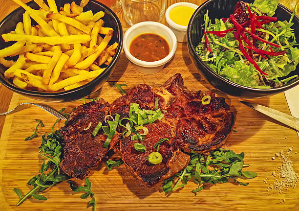 Closeup hearty meal plate with steak chop on a wooden board over arugula, topped with chives and flanked by bowls of golden fries and mixed salad. Small bowls of sauce and mustard complete the