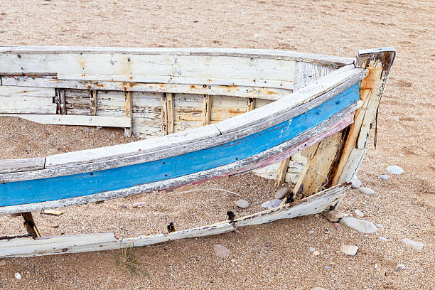 An old, weather-beaten boat rests on a sandy shore, its blue stripe peeling away. Rusty nails, rough wood, and scattered stones evoke nostalgia, solitude, and coastal history