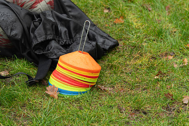 Brightly colored training cones are stacked near a black bag on the grass, surrounded by fallen leaves. It is a rainy day at the sports field, creating a vibrant scene.