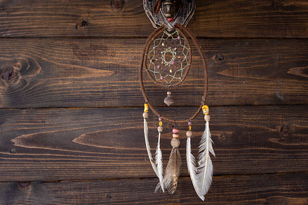 A handmade Native American style dreamcatcher with feathers and beads, hanging on a dark, textured wooden wall. A concept of rustic home decor and spiritual symbolism.