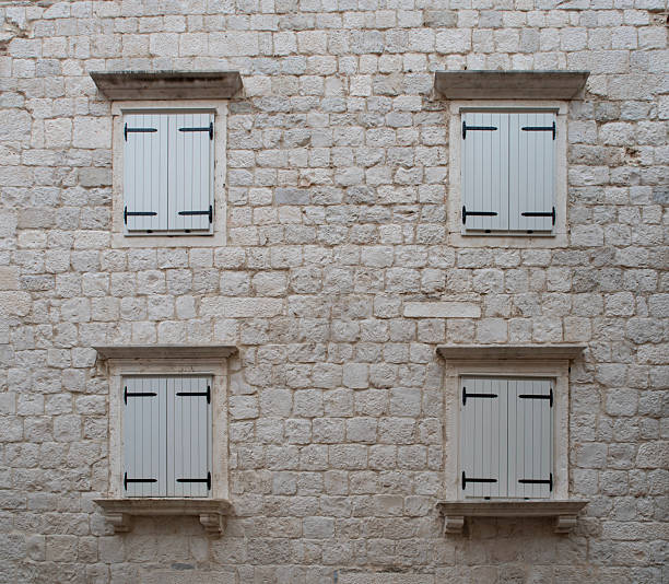 Symmetrical architectural composition featuring two arched wooden windows with metal grilles embedded in a textured stone masonry wall. Warm-toned stones create a rustic and traditional exterior design