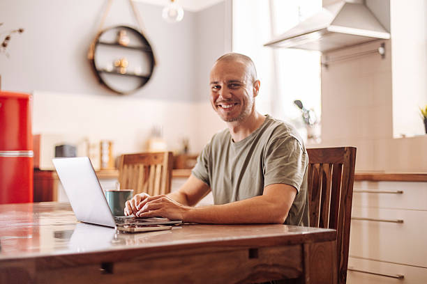 istock Man sitting at the table with a Laptop, smilling  and looing at camera while drinking coffee in the kitchen 2240622621