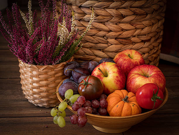 An autumn home scene, fresh fruits in a bamboo bowl, apples, plums, grapes, tomatoes and squash, heather in a basket, basket in a background, wooden floor