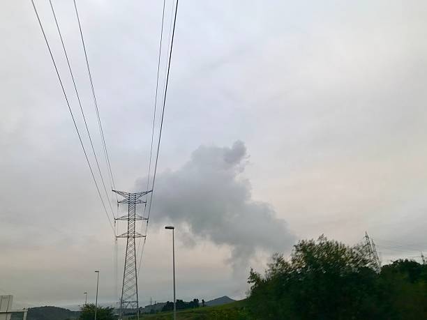 Industrial landscape with tall power lines and a cloudy sky, showing steam or emissions rising in the distance. Ideal for illustrating concepts of energy, electricity transmission, pollution, and environmental impact.