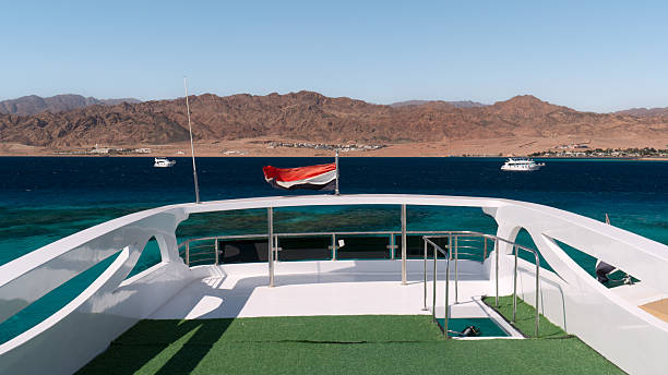 Dahab, Egypt - February 5, 2024: A view from the deck of a yacht with a flag over the Red Sea with boats in the distance, the city of Dahab and mountains on the horizon under a clear sky.