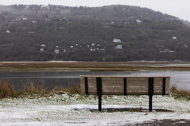 A solitary wooden bench overlooks a calm lake with snow-dusted ground and distant hillside houses, evoking quiet solitude, winter serenity, and reflective nature.