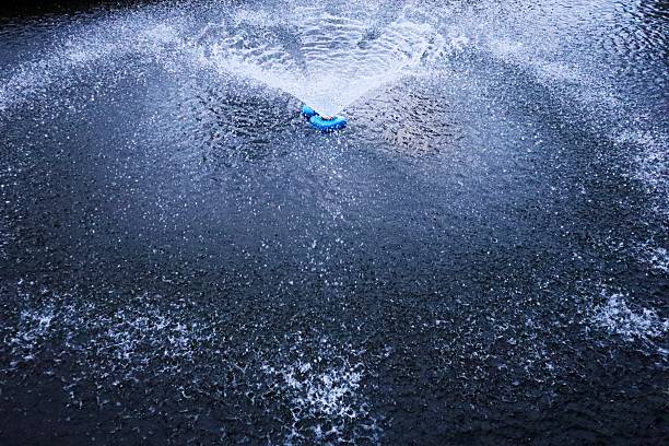 Dynamic water splash photograph capturing blue plastic bottle cap falling into dark water surface creating dramatic spray pattern. High-speed action shot showing water droplets suspended in air forming circular ripple waves around bright blue cap. The contrast between deep dark blue water and lighter splash creates striking visual effect. Perfect for concepts of impact, motion, freshness, beverage industry, recycling, environmental themes, physics of water, and action photography. Frozen moment captures energy and movement of water responding to object entry with detailed droplet formation and splash dynamics.