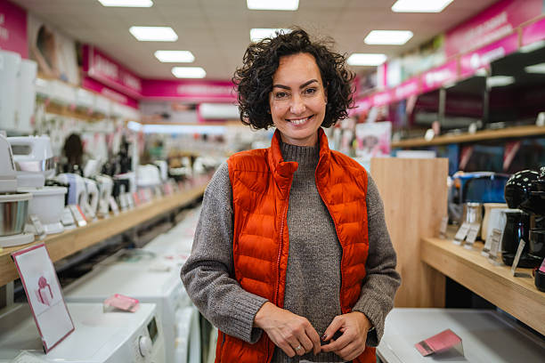 Smiling adult woman standing confidently in a modern electronics and appliance store, browsing products and enjoying a cheerful shopping experience while evaluating new household technology