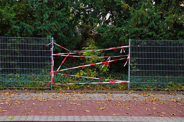 istock Metal Fence Wrapped in Red and White Caution Tape Blocking Path Amidst Green Trees and Fallen Autumn Leaves on Paved Ground 2240652442