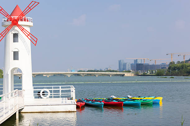 Colorful kayaks docked beside a white lighthouse on a calm urban river. Modern city skyline and construction cranes visible in the background.