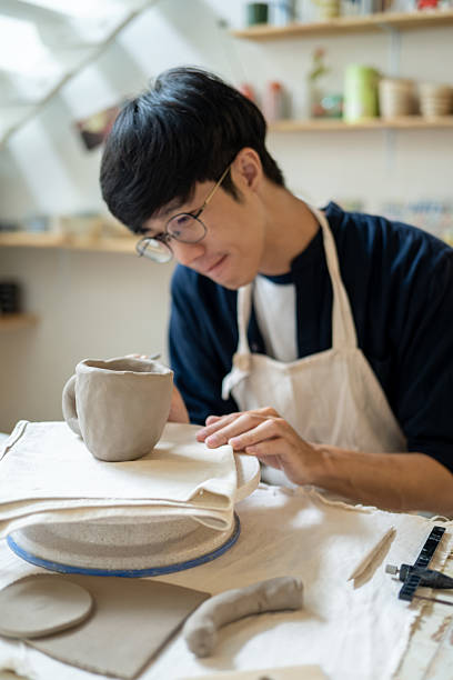 istock Young man potter artist adding texture on a clay mug at working table in a pottery class or studio. 2240684027
