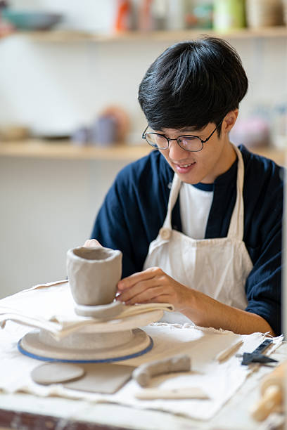 istock Young man pottery artist molding a clay mug on throwing wheel at working table in a class or studio. 2240684060