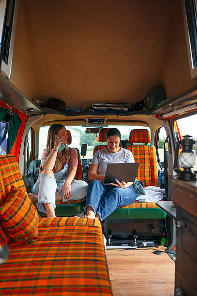 istock : Two women relaxing inside a camper van, sitting on orange plaid seats, talking and working on a laptop. 2240684666