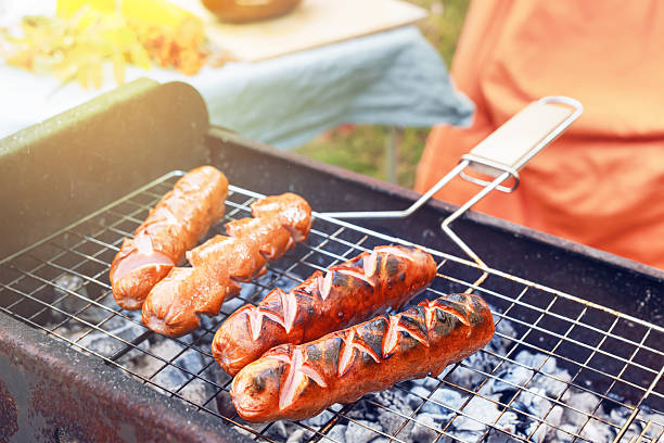 Grilled sausages are sizzling on a barbecue grill filled with glowing coals. The scene captures the essence of an autumn picnic, with warm sunlight filtering through.