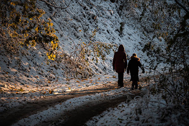 Two people walking along a snow-covered path in a winter forest, illuminated by golden light from falling sun creating long shadows