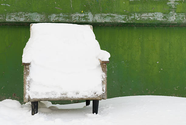 Old armchair covered with snow near a green wall in winter. Minimal urban scene with bright contrast of white snow and green background.
