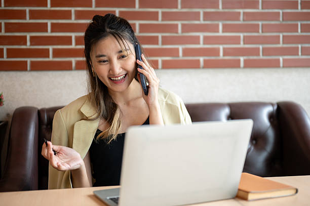 istock Asian woman holding pen as talking on phone and looking at laptop on table while sit on sofa in cafe 2240704126