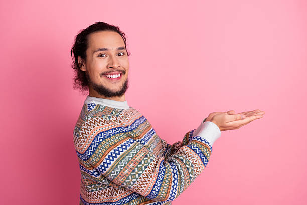 istock Happy young man in a colorful sweater posing against a pink background presenting something with an inviting hand gesture 2240706837