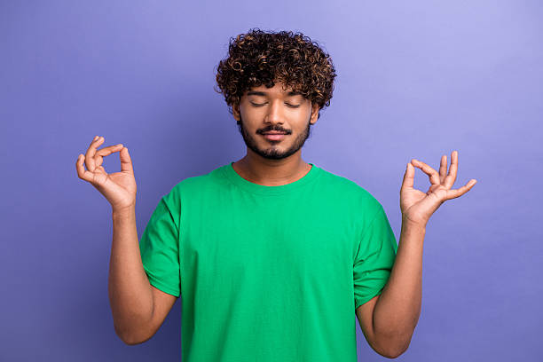 istock Relaxed young man in a green t-shirt meditating with a calm expression and hand gesture against a violet background 2240715320