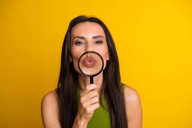istock Charming young woman with a magnifying glass emphasizing her lips in front of a vibrant yellow studio backdrop. 2240737509