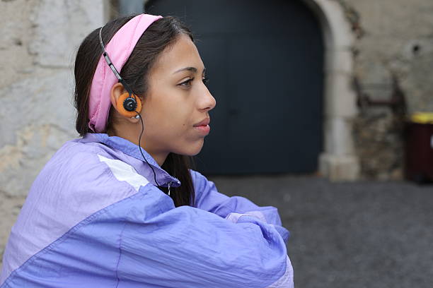 A very cute Hispanic teenage girl is listening to music with her orange vintage headphones from the 1980s. She wears a purple, pink and white cool urban outfit.
