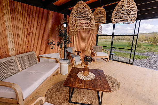 istock Scandinavian-style wooden lounge with wicker pendant lights, cozy seating, rustic table, and panoramic glass wall opening to a peaceful natural landscape. 2240749799