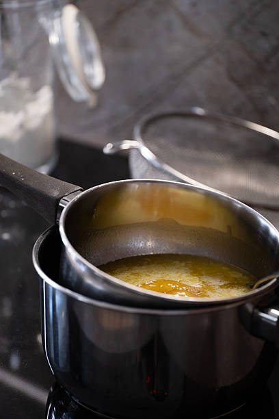 Smooth, fully melted butter in a saucepan over a double boiler setup on a black stovetop, with a spoon partially visible for stirring. This closeup kitchen image is ideal for illustrating cooking techniques, baking steps, food prep visuals, and recipe instructions for culinary blogs or educational materials.