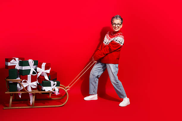 istock Festive woman pulling a gift-laden sleigh in cozy red sweater on bright red background, spreading holiday joy 2240750128