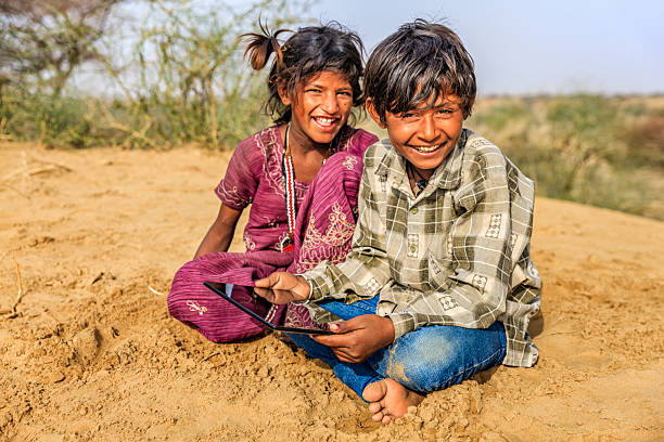 Happy Indian children sitting on a sand dune and using digital tablet in desert village, Thar Desert, Rajasthan, India.