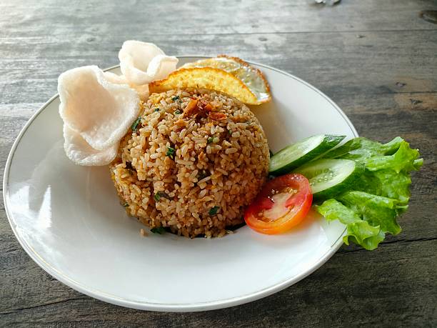 a neatly plated portion of Indonesian fried rice, or nasi goreng, arranged in a round shape on a white dish. The rice is mixed with spices, vegetables, and bits of egg, giving it a rich brown color. On the side, there are fresh cucumber slices, a tomato slice, and a piece of lettuce for garnish, adding vibrant green and red accents to the plate. A sunny-side-up fried egg is placed behind the rice, along with several light, crispy crackers. The wooden table beneath the plate creates a rustic dining atmosphere, making the dish look warm, inviting, and appetizing.