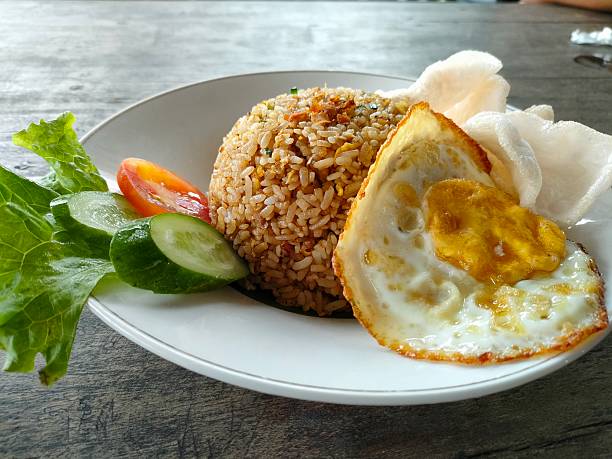 a plate of Indonesian fried rice served neatly in a dome shape, accompanied by a sunny-side-up egg placed at the front of the dish. Fresh vegetables, including slices of cucumber, a piece of tomato, and a leaf of lettuce, are arranged on the side, adding a vibrant touch to the presentation. A few crispy prawn crackers rest behind the rice, completing the traditional serving style. The dish sits on a rustic wooden table, and the overall scene highlights a warm, appetizing meal that looks both simple and satisfying.