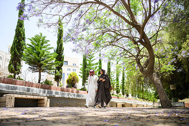 istock Wide shot of Middle Eastern couple walking together along a scenic tree lined pathway with purple blooms in a park 2240772107