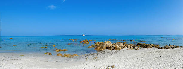 Bright sunny day with clear blue sea and rocky coastline. Sailboat anchored near shore with green hills in the background. Scenic coastal landscape with calm ocean water and natural rocks.