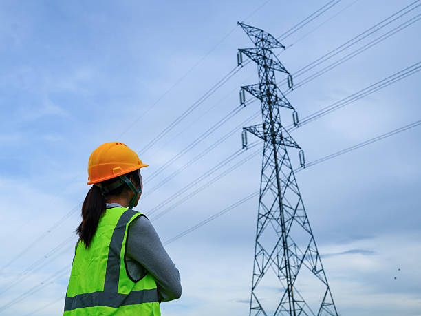 istock Engineer in Hard Hat and Safety Vest Observing High Voltage Power Lines Against Blue Sky with Clouds 2240776680