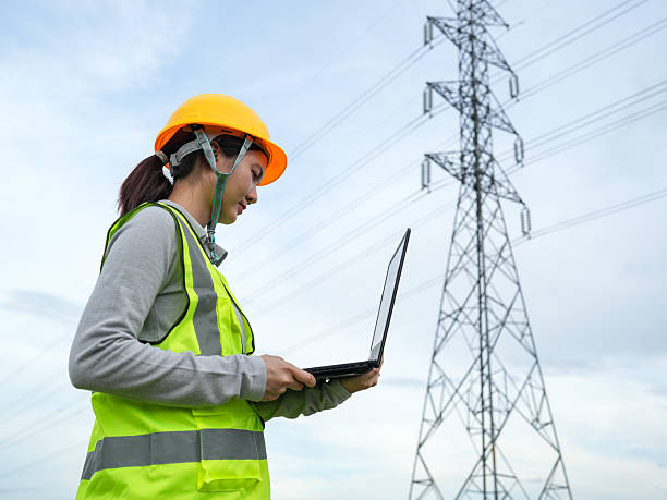istock Female Engineer with Hard Hat and Laptop Conducting Field Work near Electrical Transmission Tower in Bright Yellow Safety Vest Outdoors 2240776784