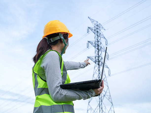 istock Female engineer examines power lines while using laptop in outdoor electrical site, wearing safety gear for fieldwork, under clear blue sky with clouds 2240776797