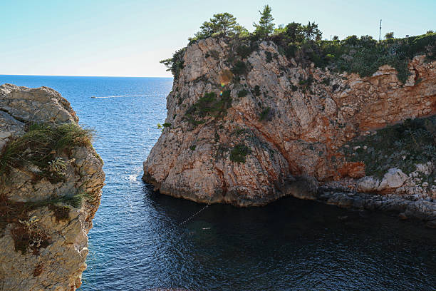 A serene view of rugged coastal cliffs meeting the calm blue waters of the Mediterranean Sea. The sunlight highlights the textured rock formations and lush greenery, creating a peaceful natural scene perfect for travel and landscape photography collections.