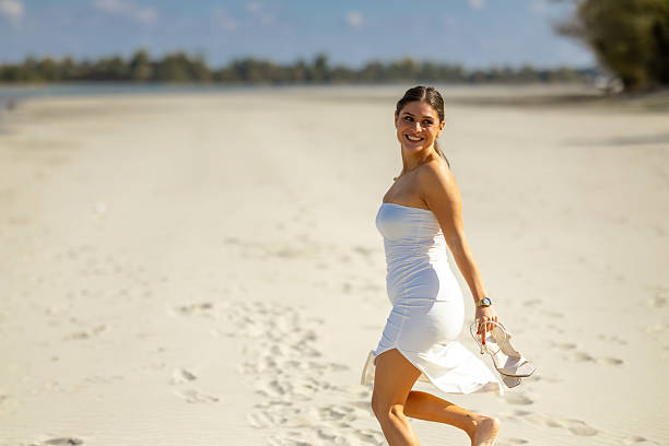 A girl in a white dress and shoes in her hands runs happily on the beach and turns towards the camera.