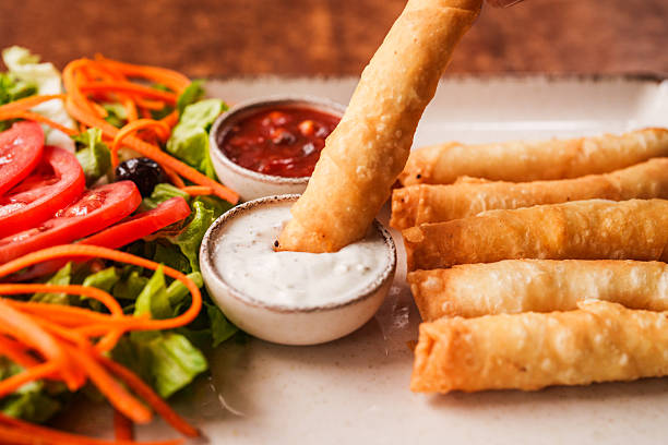 A hand dips a crispy Turkish sigara böreği into a creamy yogurt-based dip, surrounded by a vibrant fresh salad and neatly arranged cheese rolls. A delicious bite in the making.
